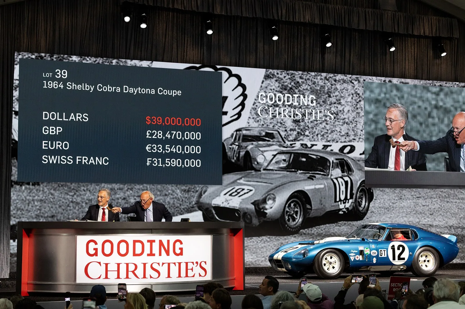 a photograph of an auction, we can see bids being taken, beside is a vintage blue racecar that is being sold