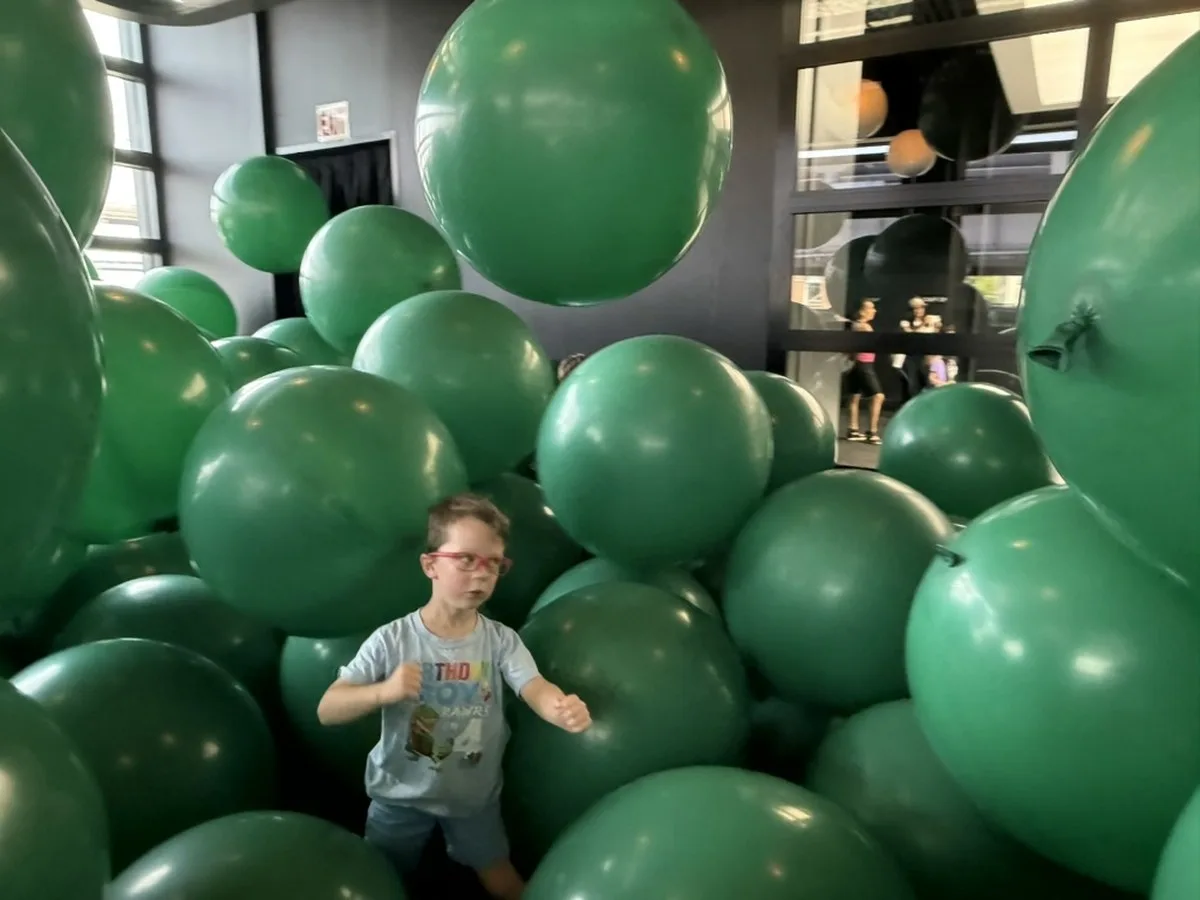 Installation view of Martin Creed's "Work No. 3883: Half the air in a given space" at the Balloon Museum, filling a room with green balloons; a young boy stands in the foreground among them, with onlookers visible through a window behind.