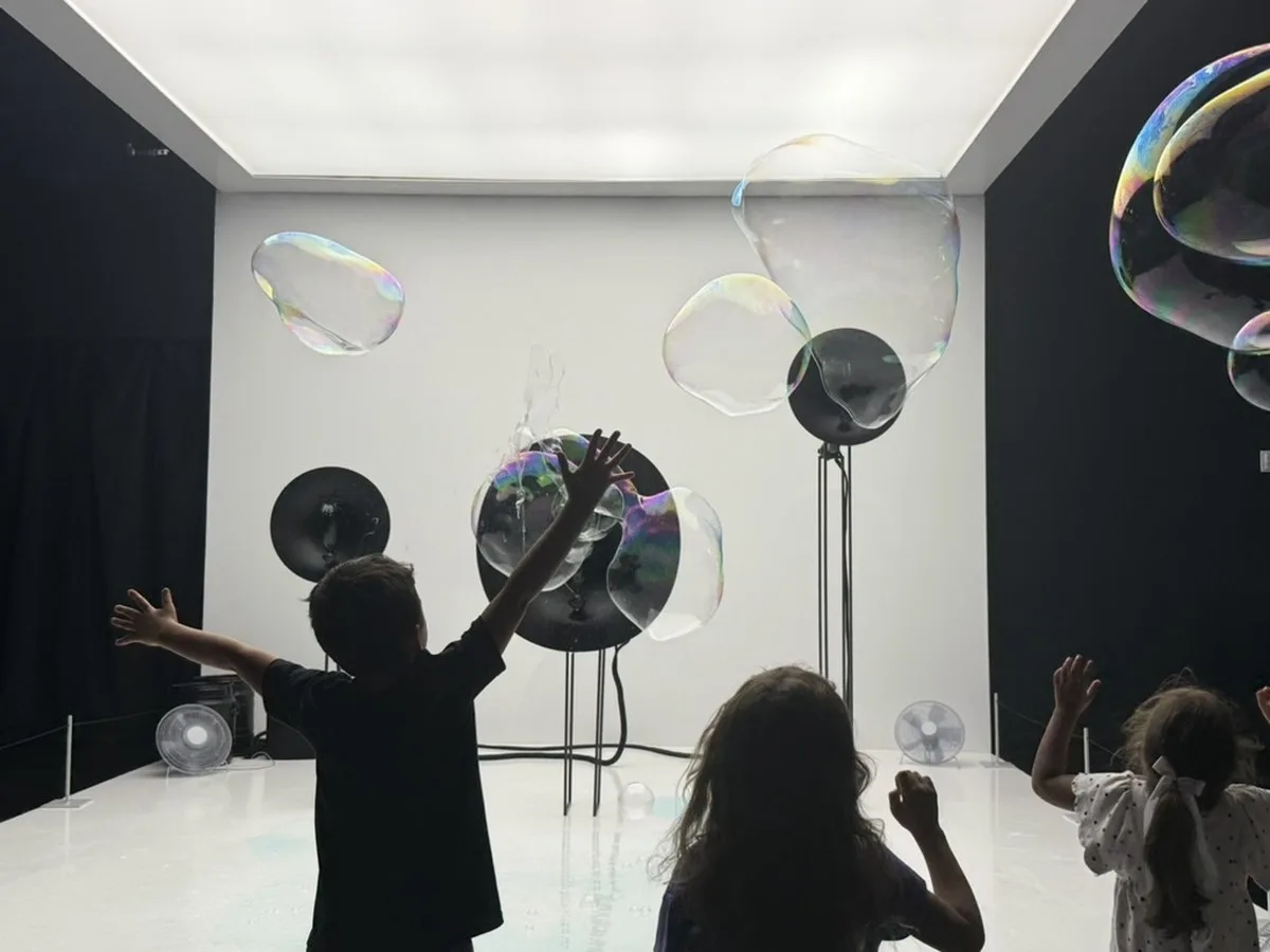 Children stand with arms raised in a dark, minimalist room, reaching toward large iridescent soap bubbles generated by fan-and-ring devices mounted on stands against a white wall.