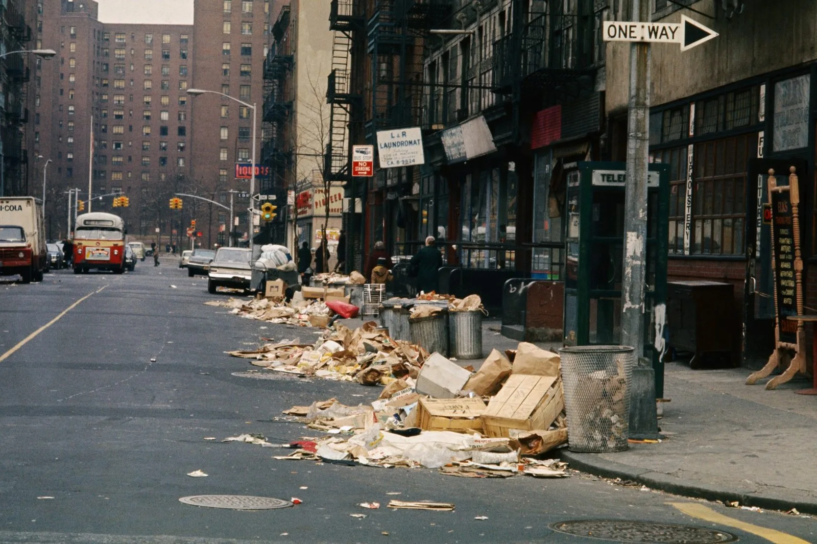Archival color photograph of a New York City street lined with large trash piles along the curb. In the foreground, uncollected garbage — flattened cardboard boxes, torn paper bags, wooden crates, and loose debris — spills from overflowing metal trash cans onto the sidewalk and into the gutter for most of a block. A green telephone booth marked "TELEPHONE" and a "ONE WAY" street sign stand near the curb. Behind the trash, a row of storefronts includes "L & R LAUNDROMAT" and a furniture/upholstery shop, with tenement buildings and fire escapes rising above. A "BUS STOP NO STANDING" sign and a "LIQUORS" sign are visible further down the block. A vintage red-and-cream city bus, a boxy delivery truck, and several parked cars line the street, with a few pedestrians walking near the shops. In the far background, a tall brick high-rise apartment building overlooks the scene under an overcast sky.