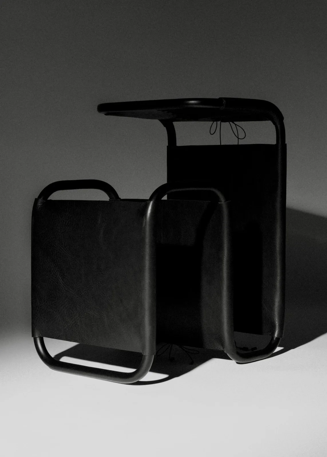 A side table made with a black metal frame. The metal bends back on itself several times. It has taught black leather wrapped around several parts of the frame, tied together with black chord to imitate a corset.