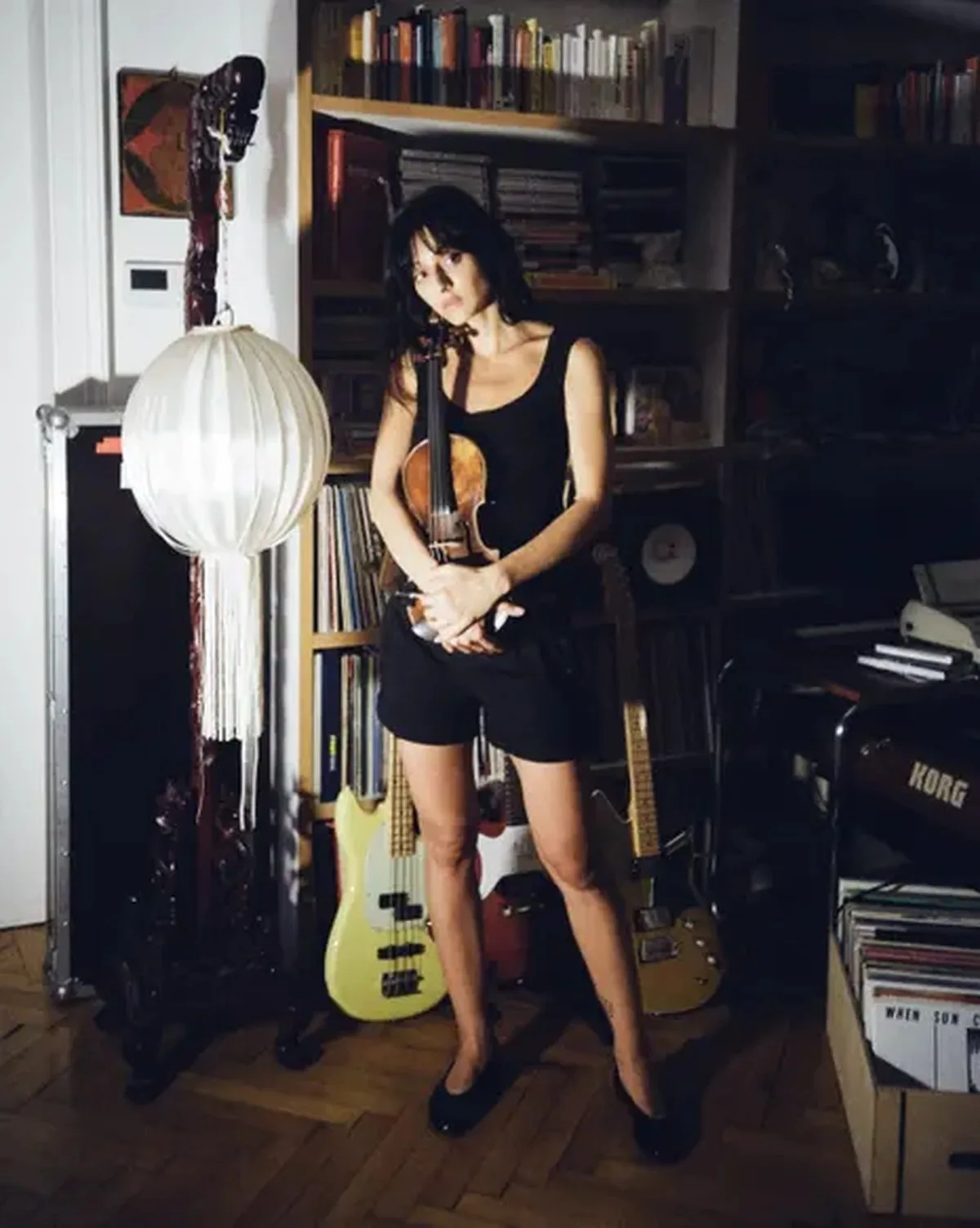 Singer Sofie Royer holds a violin, standing before a bookshelf and paper lantern lamp