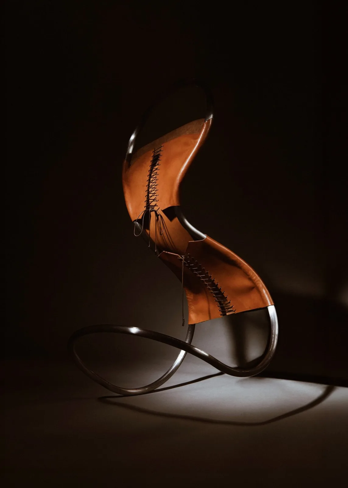 A chair made from a continuous, tubular, stainless steel frame covered with tanned leather and secured with brown cord that has been laced like a corset. The chair is leant forward and to the left and looks like it is precariously balanced.
