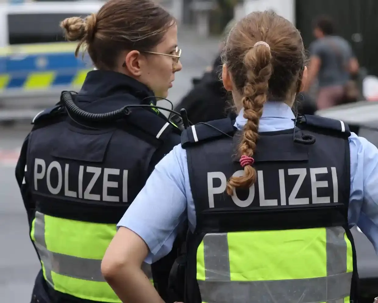 Two German police officers wearing "Polizei" vests stand together at a cordoned-off scene
